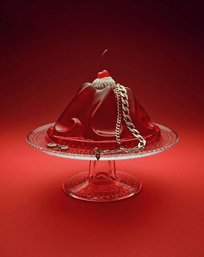 Red gelatin dessert with a cherry on top and gold chain on a clear pedestal cake stand against a matching red background.