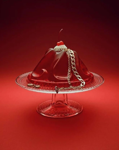 Red gelatin dessert with a cherry on top and gold chain on a clear pedestal cake stand against a matching red background.