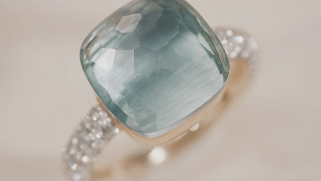 Close-up of a ring with a large, faceted, square-cut, light-blue gemstone, and a gold band with embedded diamonds. Jewellery advertisement.