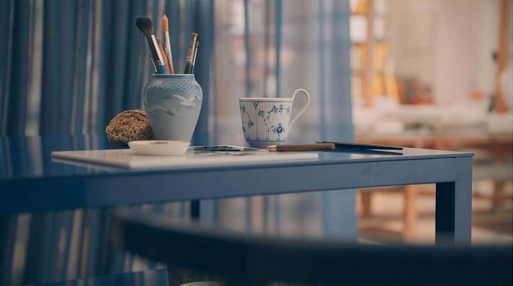 Still life on a blue table includes a vase with art brushes, sponge, teacup, writing pen, and eyeglasses in a blurred room setting.