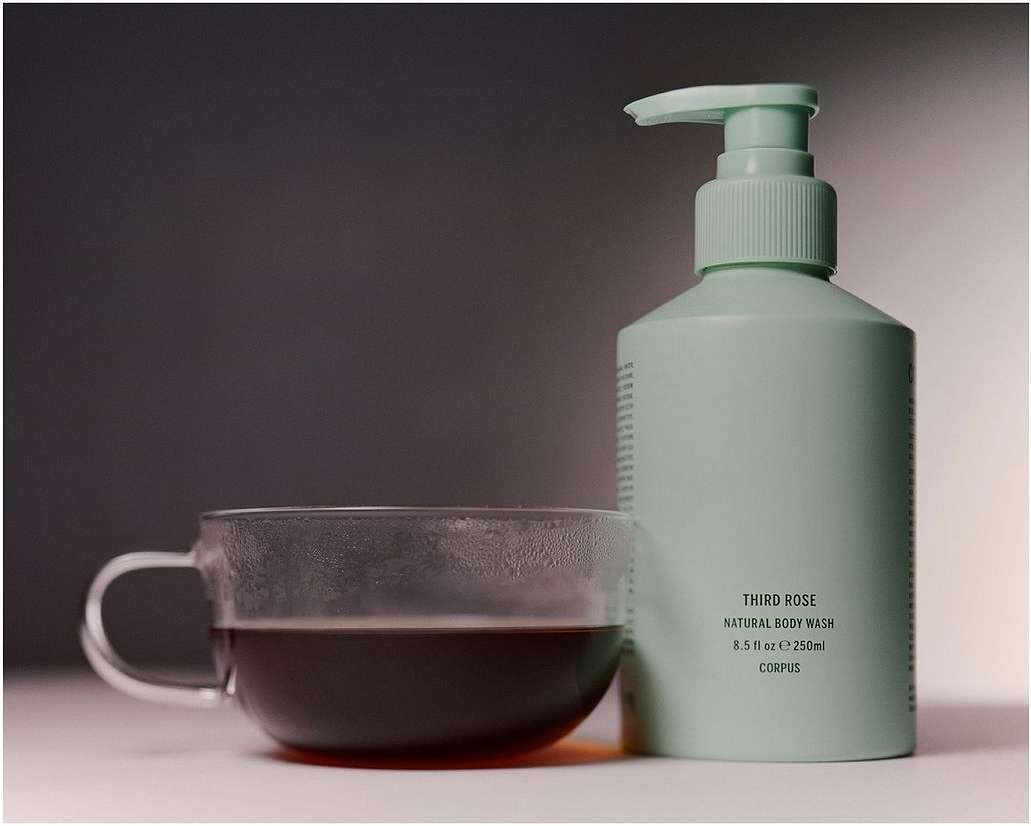 Minimalist product shot featuring a glass coffee cup and a mint-green bottle of body wash against a soft, graduated grey background.