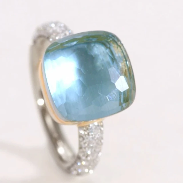 Cushion-cut blue gemstone ring featuring a band studded with pavé diamonds and set against a neutral background. Jewelry advertisement.