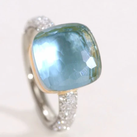 Cushion-cut blue gemstone ring featuring a band studded with pavé diamonds and set against a neutral background. Jewelry advertisement.
