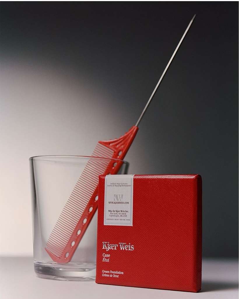 Still life photograph of a red Kjaer Weis makeup compact, a red YS Park comb in a clear glass, against a grey backdrop.
