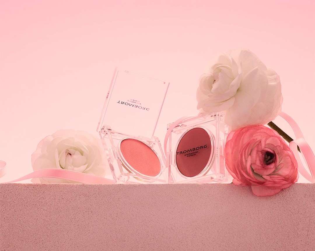 Two Tromborg blush compacts stand beside pink and white ranunculus flowers, displayed against a soft pink backdrop for a beauty still life.
