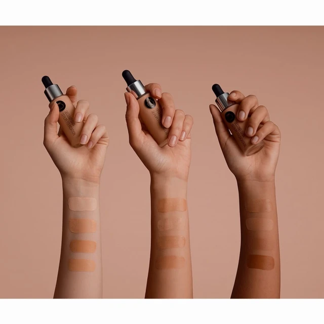 Three hands displaying swatches and holding foundation bottles with droppers against a beige background in a beauty product shot.