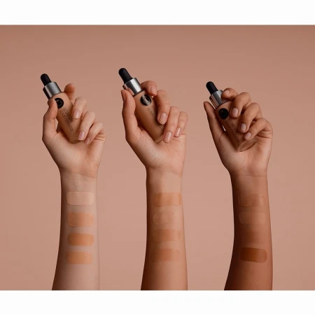 Three hands displaying swatches and holding foundation bottles with droppers against a beige background in a beauty product shot.