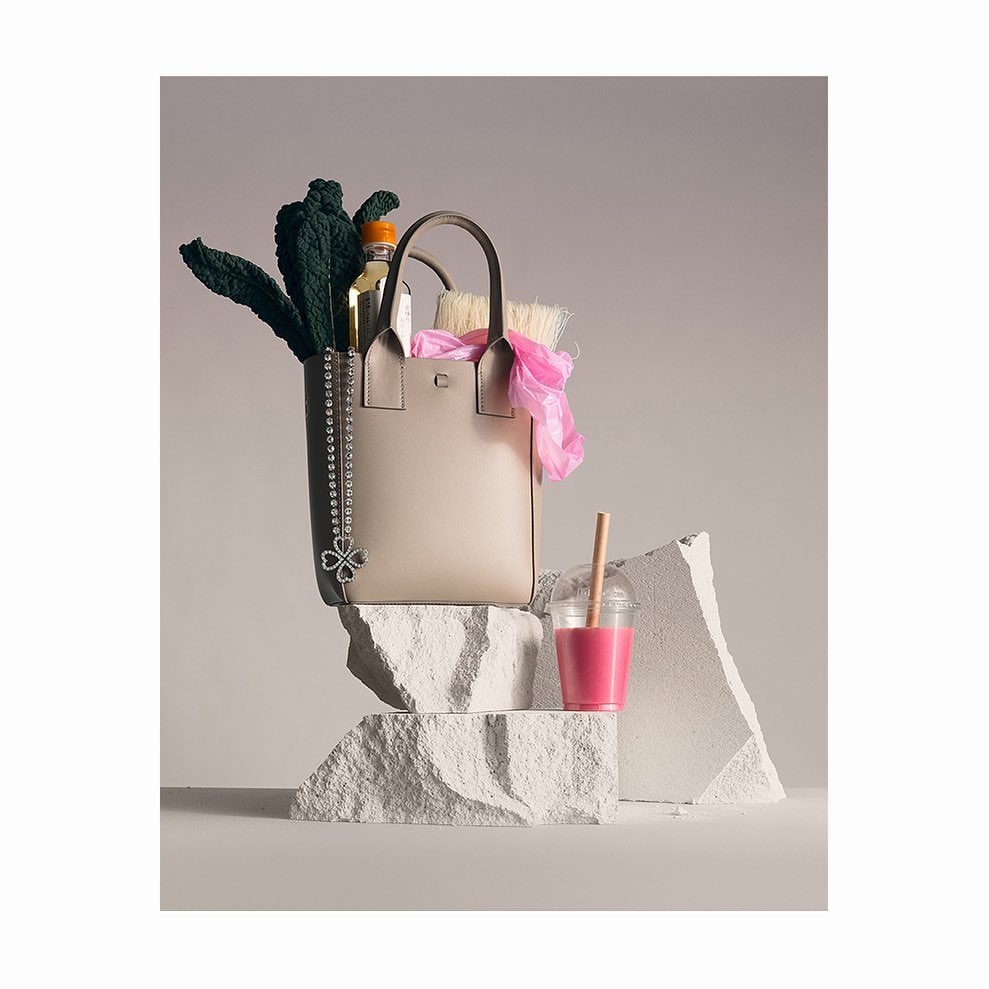 Beige leather tote bag with a sparkly chain, filled with produce and accessories, sits next to a pink drink on rock slabs.