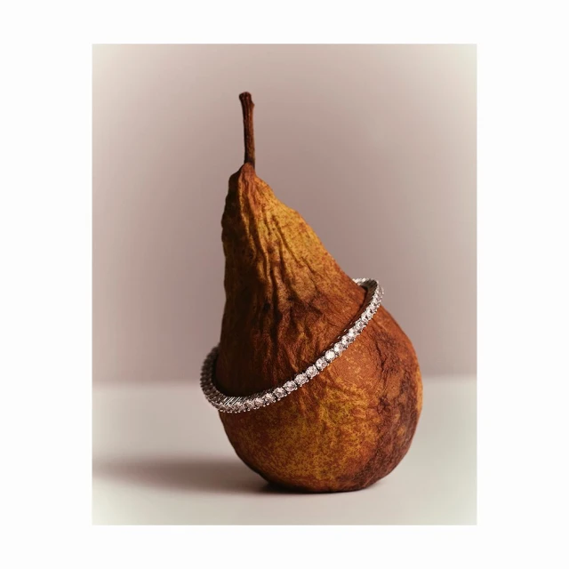 A withered pear is adorned with a diamond bracelet, set against a soft neutral backdrop, with studio lighting highlighting its textures.