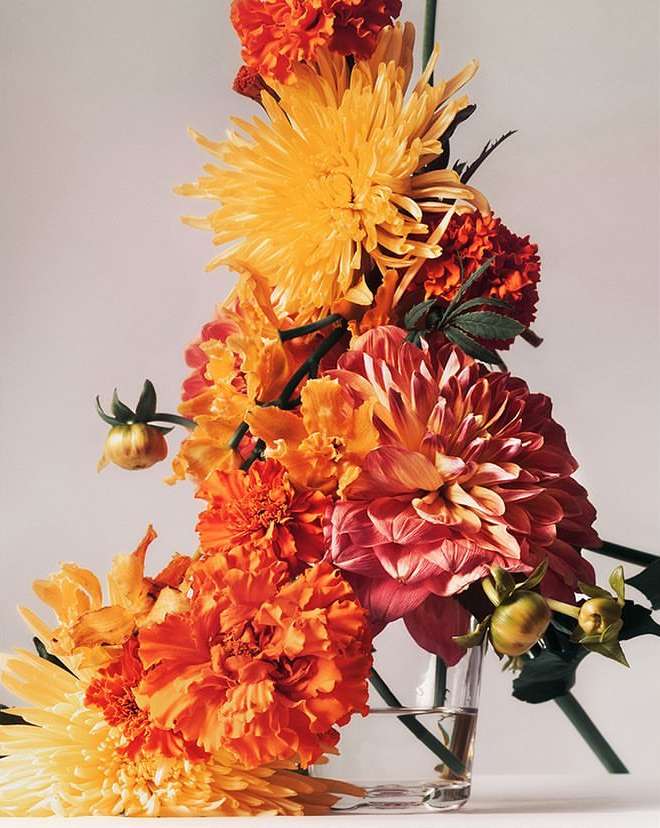 Floral arrangement featuring vibrant orange and yellow chrysanthemums and dahlias in a clear glass vase against a pale backdrop.