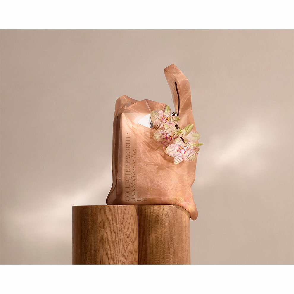 Tan-colored tote bag made of sheer fabric, sitting on light wood pedestals, adorned with orchid flowers, lit softly against a neutral backdrop.