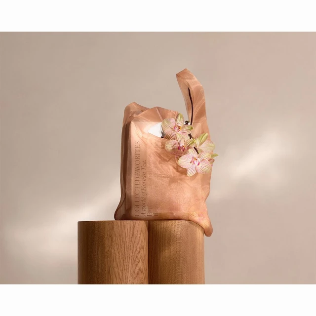 Tan-colored tote bag made of sheer fabric, sitting on light wood pedestals, adorned with orchid flowers, lit softly against a neutral backdrop.