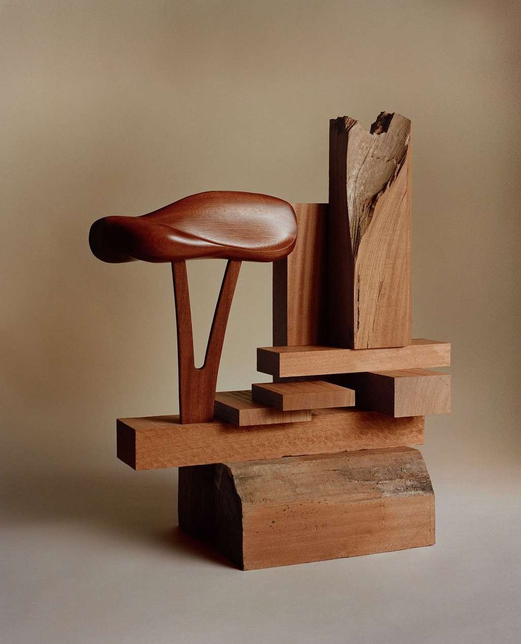 Abstract sculpture featuring stacked wooden blocks and a carved wooden seat atop a light tan background in a studio setting.
