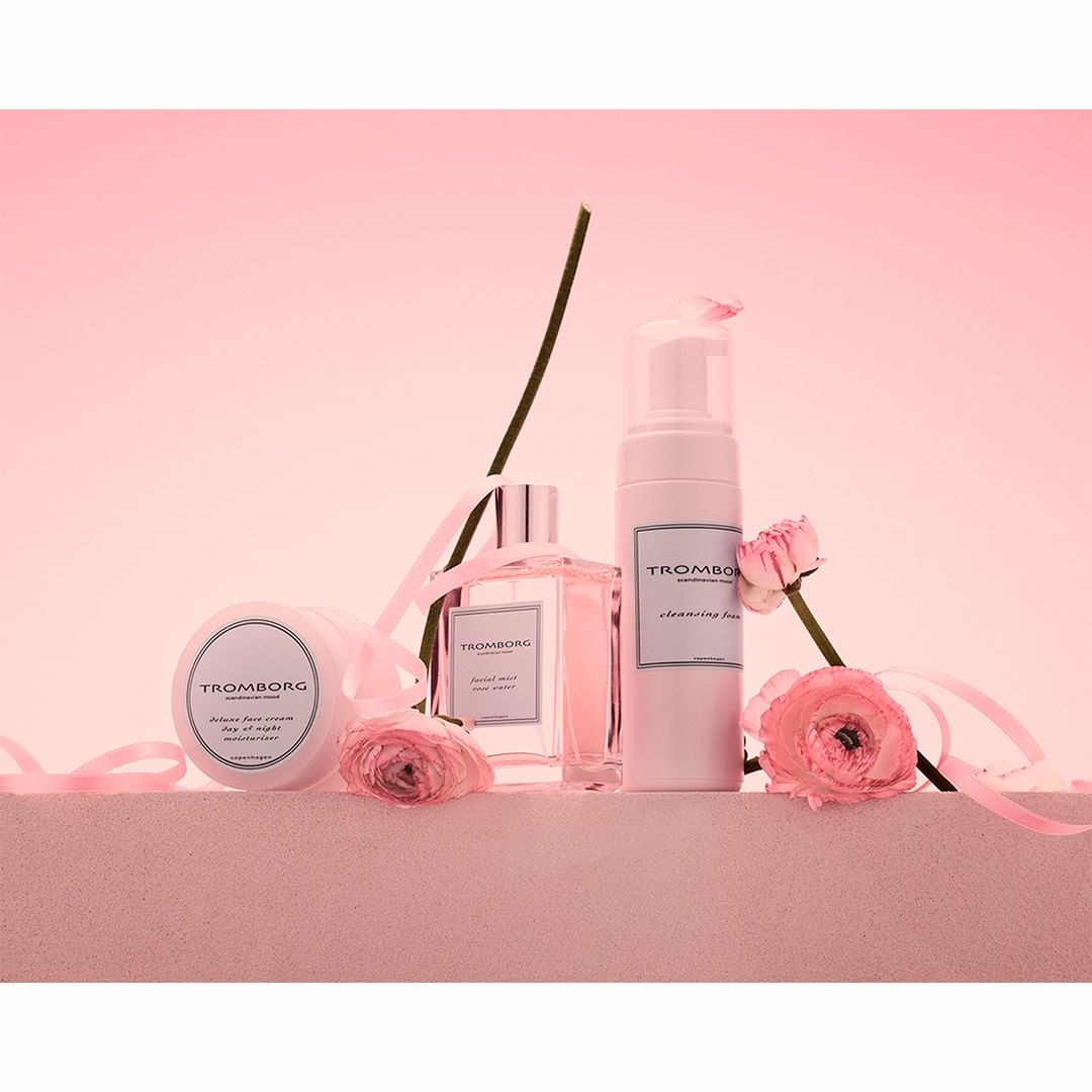 Still life shot of Tromborg beauty products, including cleanser, perfume and body cream, arranged with pink ribbons and flowers.