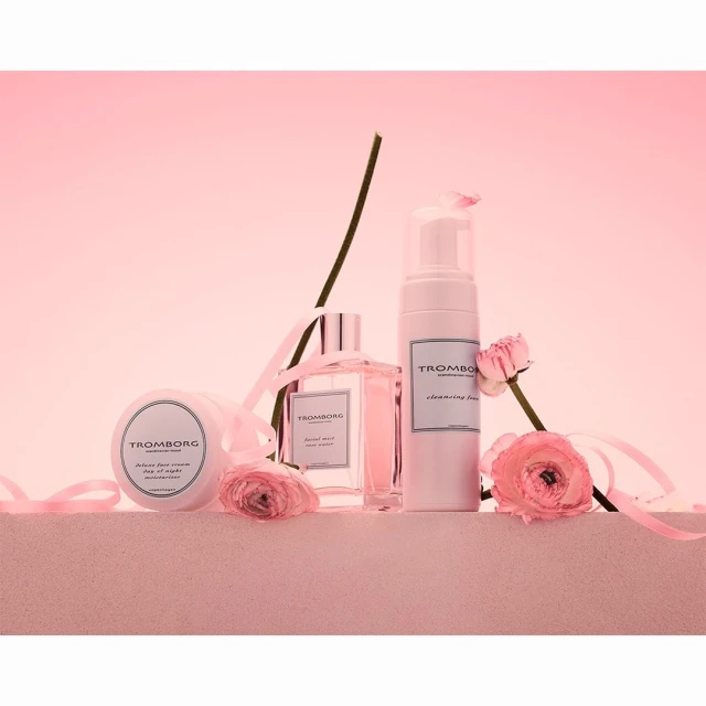 Still life shot of Tromborg beauty products, including cleanser, perfume and body cream, arranged with pink ribbons and flowers.