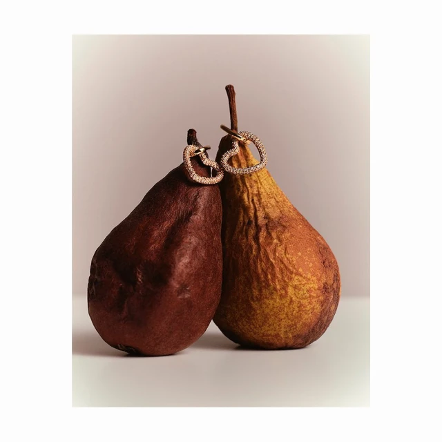 Still life shot of two pears, one brown and one yellow, with heart-shaped jewelry pieces attached, set against a neutral background.