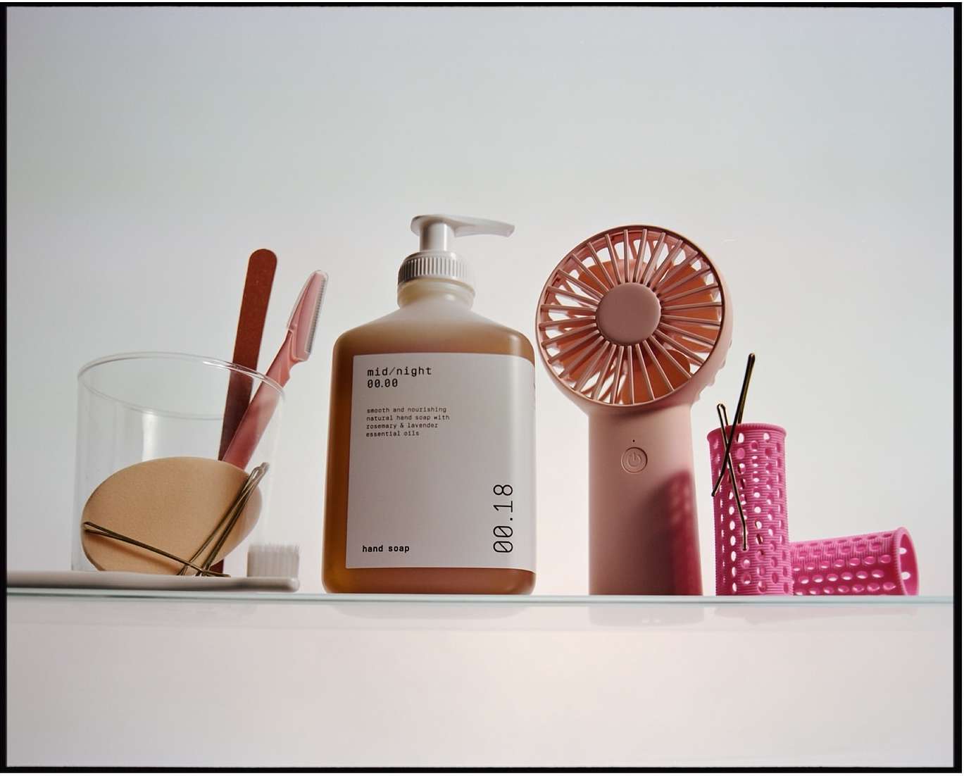 Still life of a soap dispenser, personal fan, hair curlers, and skincare items in pastel shades, arranged on a glass shelf against a white background.