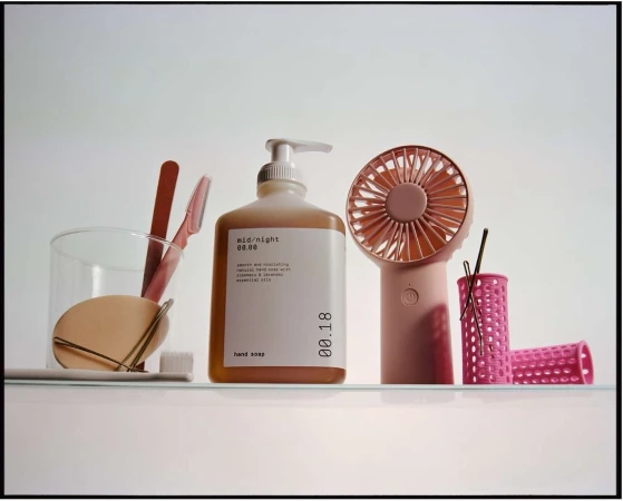 Still life of a soap dispenser, personal fan, hair curlers, and skincare items in pastel shades, arranged on a glass shelf against a white background.