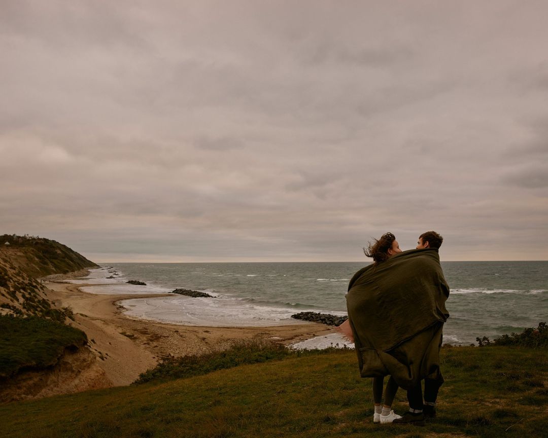 A young couple stands wrapped in a green blanket on a grassy hill overlooking the ocean under a cloudy sky in a romantic outdoor scene.