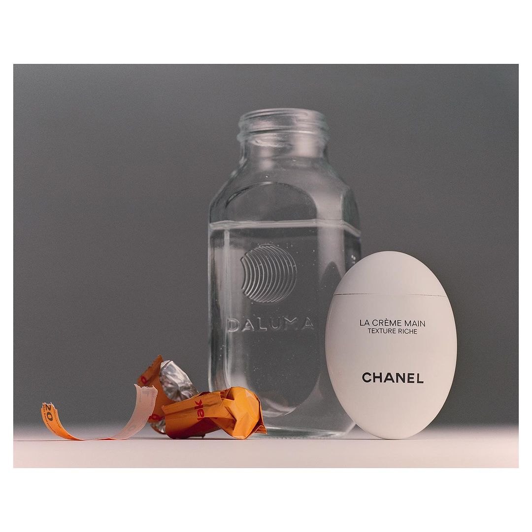 Still life featuring a clear bottle, a white Chanel cream container, and a foil-wrapped candy on a white surface against a gray backdrop.