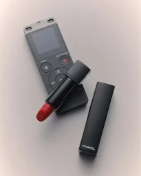 An open red lipstick and its black case resting on top of a digital voice recorder against a neutral grey background