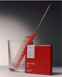 Red Kjaer Weis cosmetic case placed next to a clear glass and a red tail comb against a gradient background