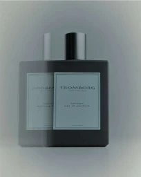A dark glass perfume bottle with a black cap and pale label set against a minimalist gradient grey studio background.