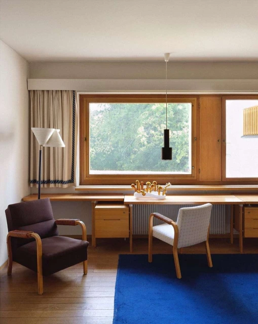 Interior view of room with a brown chair, a gray chair, a wooden desk, blue rug, curtains and natural light from window.