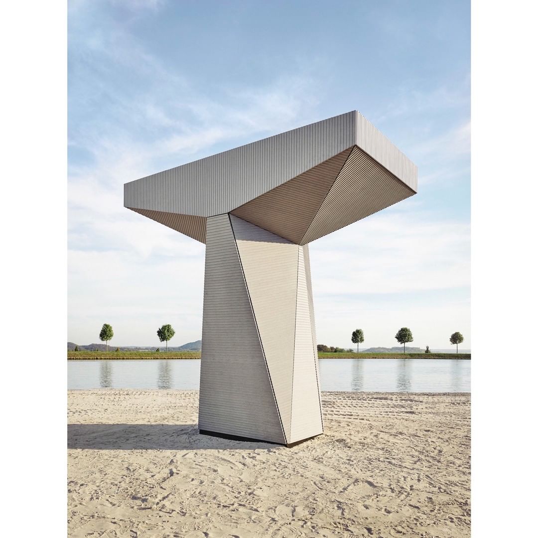 Modern architectural sculpture of a geometric observation point on a sandy beach with distant trees and a blue sky.
