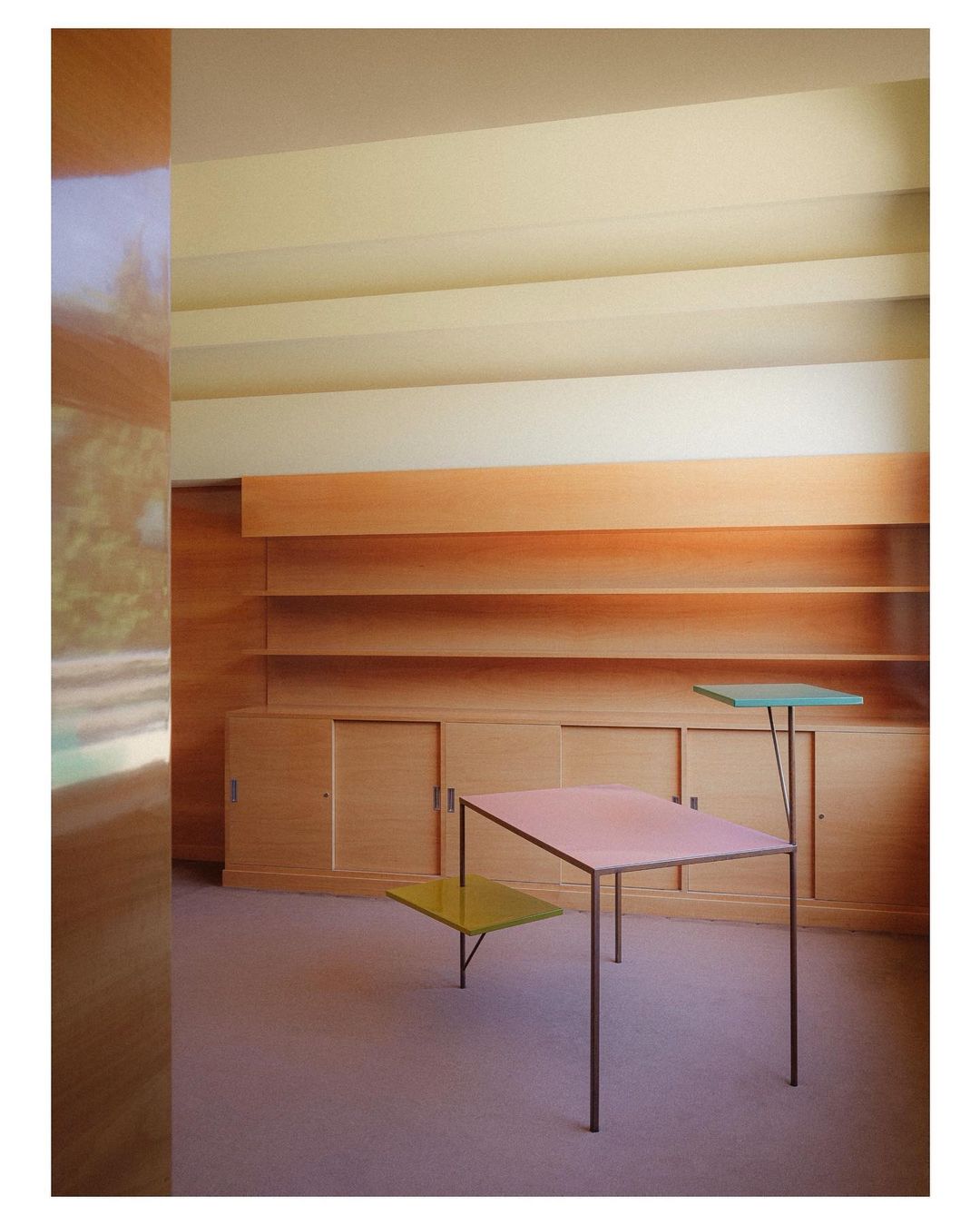 Architectural interior showcases a modern table with multiple colored surfaces, wooden shelving, and a minimalist design aesthetic.