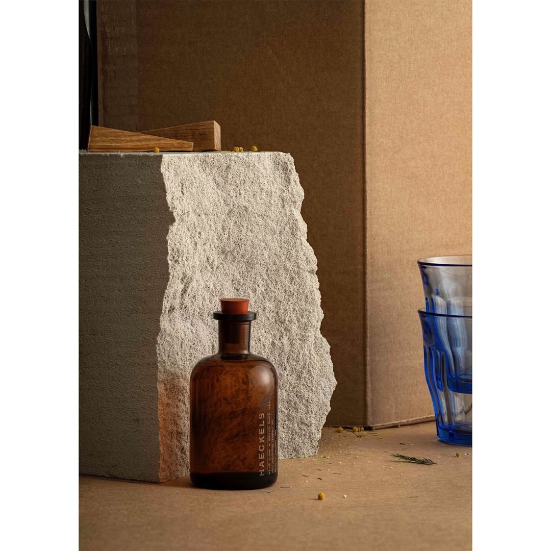 Still life featuring an amber glass bottle with a cork stopper, set against a light stone slab and cardboard boxes with blue glassware.