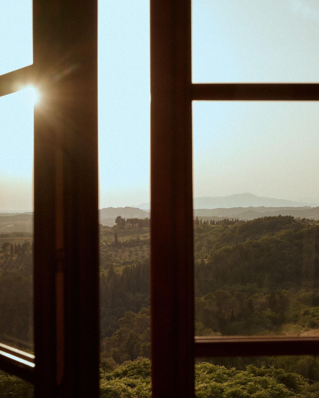 Open window view of lush green valley with soft sunlight and distant hills under a clear sky, creating a serene landscape scene.