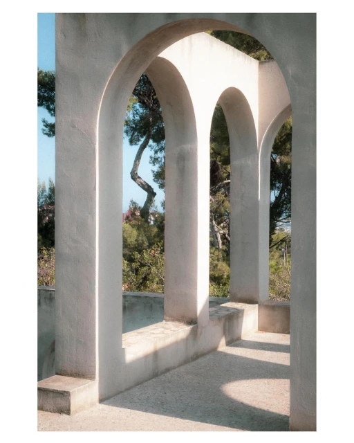 Architectural photograph of arched concrete hallway, light and shadows on floor, greenery and blue sky visible through openings.