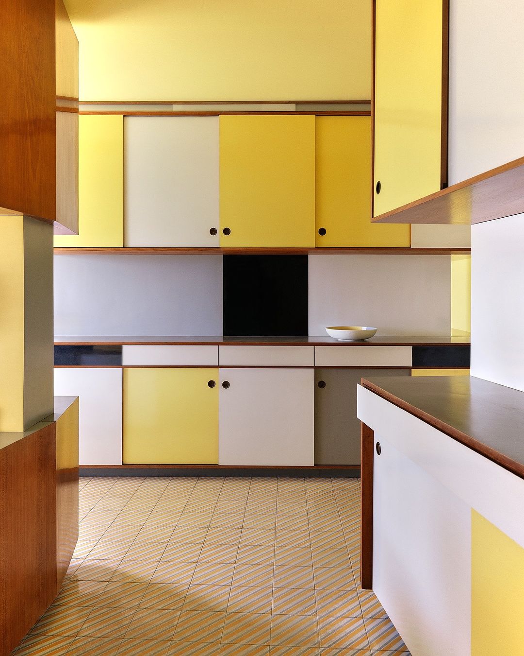 Modern kitchen design featuring a geometric layout with yellow, white, black and wooden cabinets, complemented by a patterned tile floor.