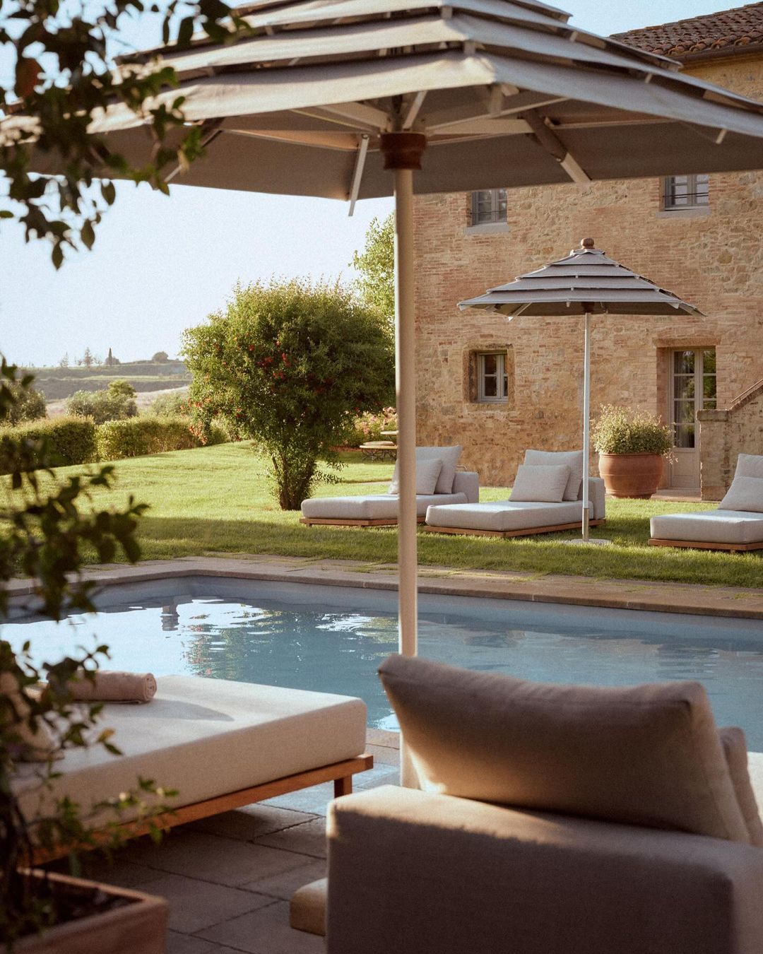 Outdoor lounge area with a swimming pool, featuring sun loungers, umbrellas, and a stone building in a grassy environment on a bright day.