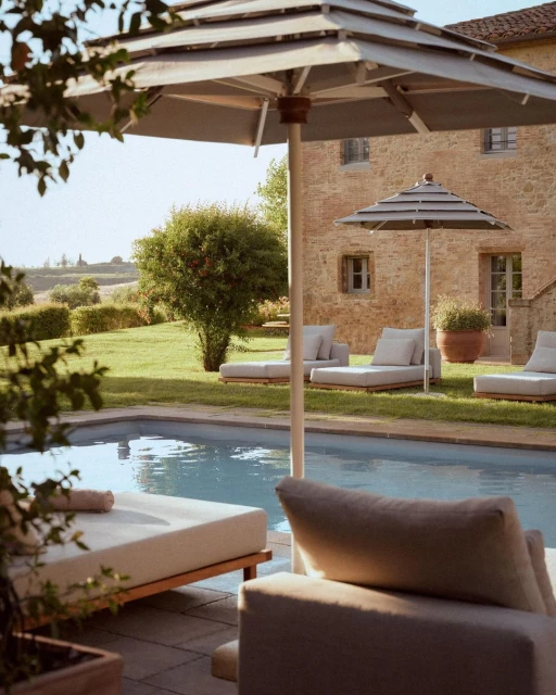Outdoor lounge area with a swimming pool, featuring sun loungers, umbrellas, and a stone building in a grassy environment on a bright day.
