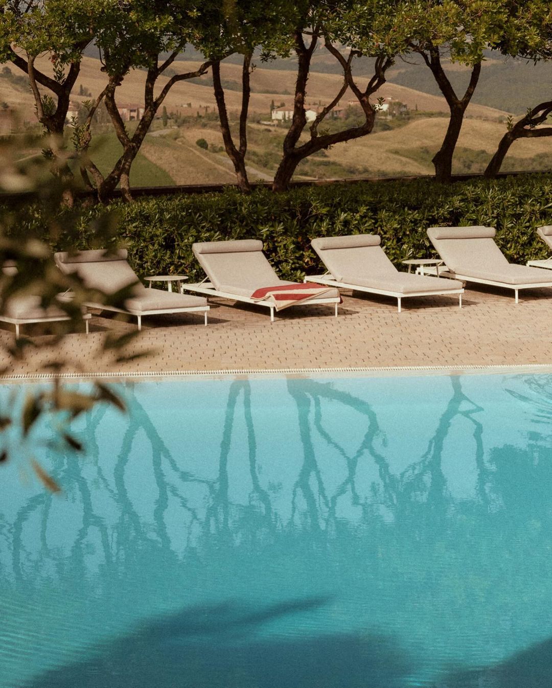 Swimming pool scene with deck chairs, trees, and a distant villa on a sunny day, offering a relaxing and luxurious resort experience.