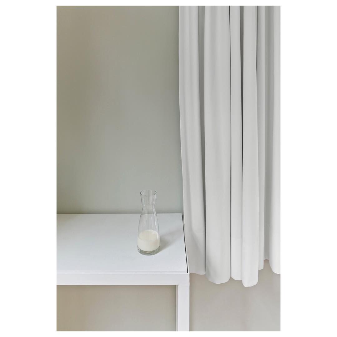 A glass milk bottle sits on a white table against a grey wall and white curtain backdrop in a naturally lit, minimalist setting.