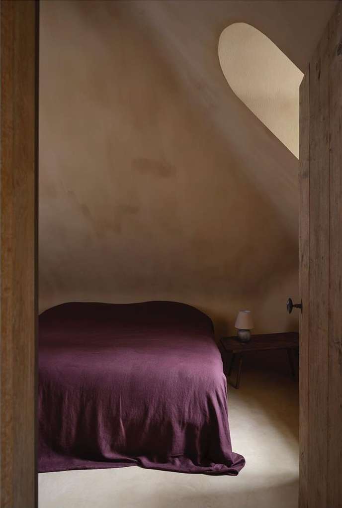 Interior shot of a bedroom in an attic with a bed covered in a dark purple blanket, visible through an open wooden door.