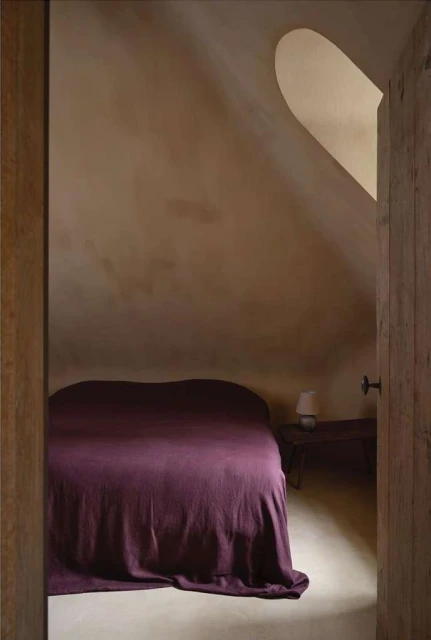 Interior shot of a bedroom in an attic with a bed covered in a dark purple blanket, visible through an open wooden door.