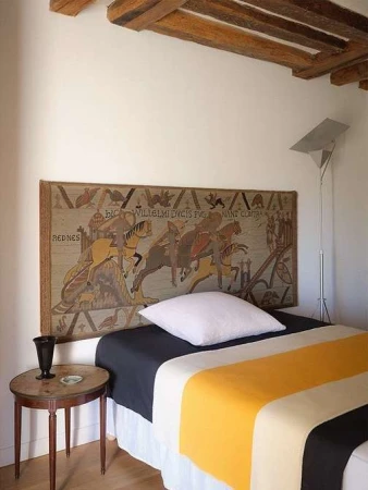 Bedroom interior with a tapestry headboard depicting a historic scene, a color-blocked bedspread, and a vintage table with a black vase.