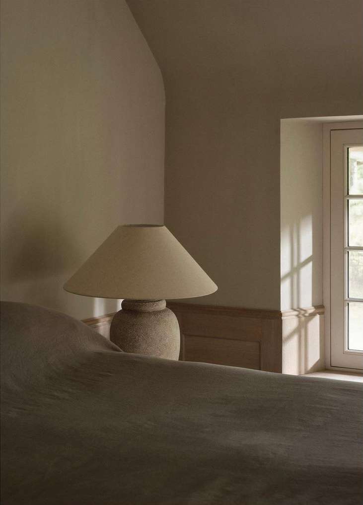 Bedroom interior with a table lamp on a bedside table, window with light streaming in, creating a soft, warm atmosphere.