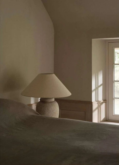 Bedroom interior with a table lamp on a bedside table, window with light streaming in, creating a soft, warm atmosphere.
