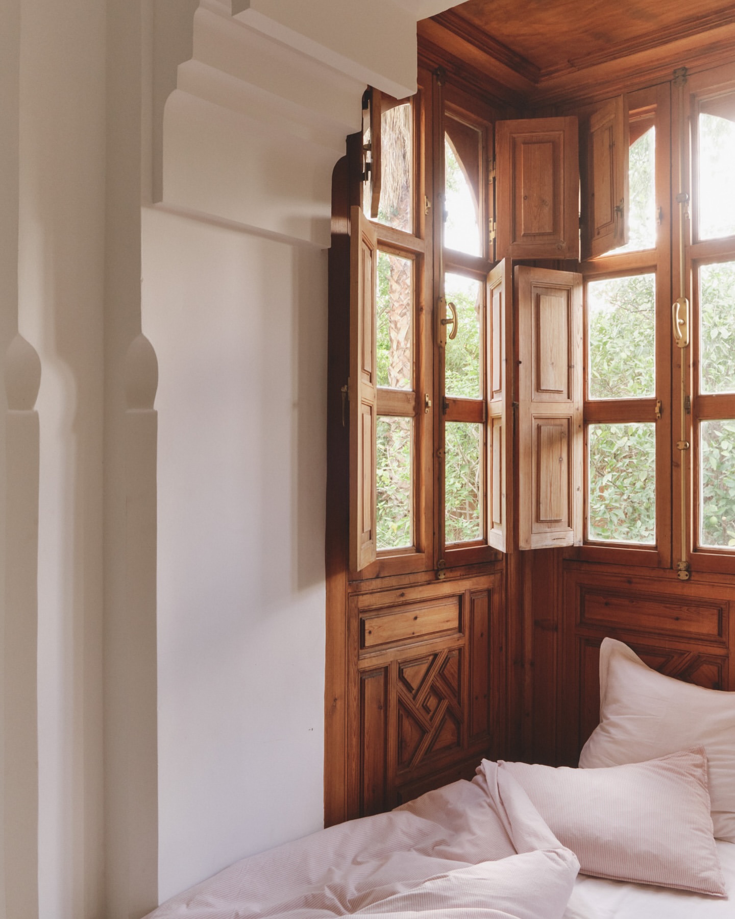 Bedroom corner with wood windows and blush bedding. Wooden details and white walls create a peaceful, relaxing ambiance. 