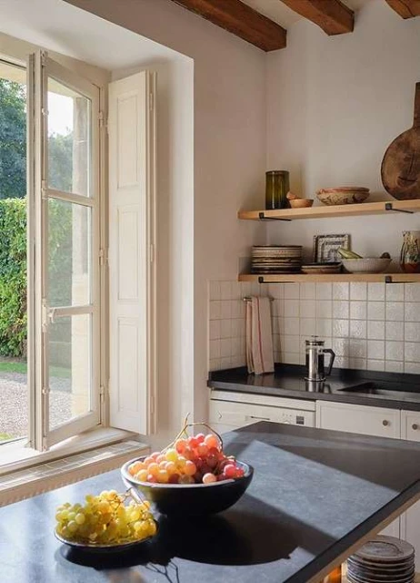 A bowl of colorful grapes on a kitchen island inside an airy, well-lit room with white walls, wooden shelves, and view of a garden.