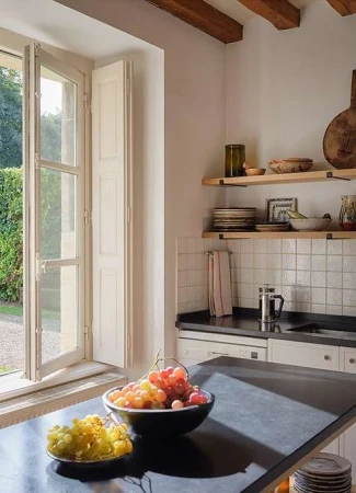 A bowl of colorful grapes on a kitchen island inside an airy, well-lit room with white walls, wooden shelves, and view of a garden.
