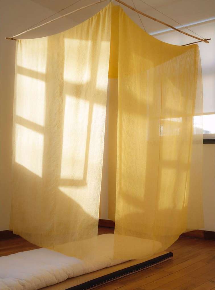 Indoor view of a semi-sheer yellow canopy or curtain hanging from a wooden rod, casting shadows on white walls and hardwood floors.