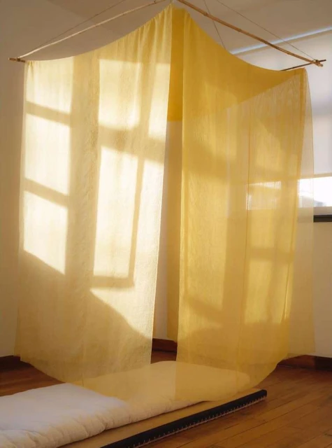 Indoor view of a semi-sheer yellow canopy or curtain hanging from a wooden rod, casting shadows on white walls and hardwood floors.