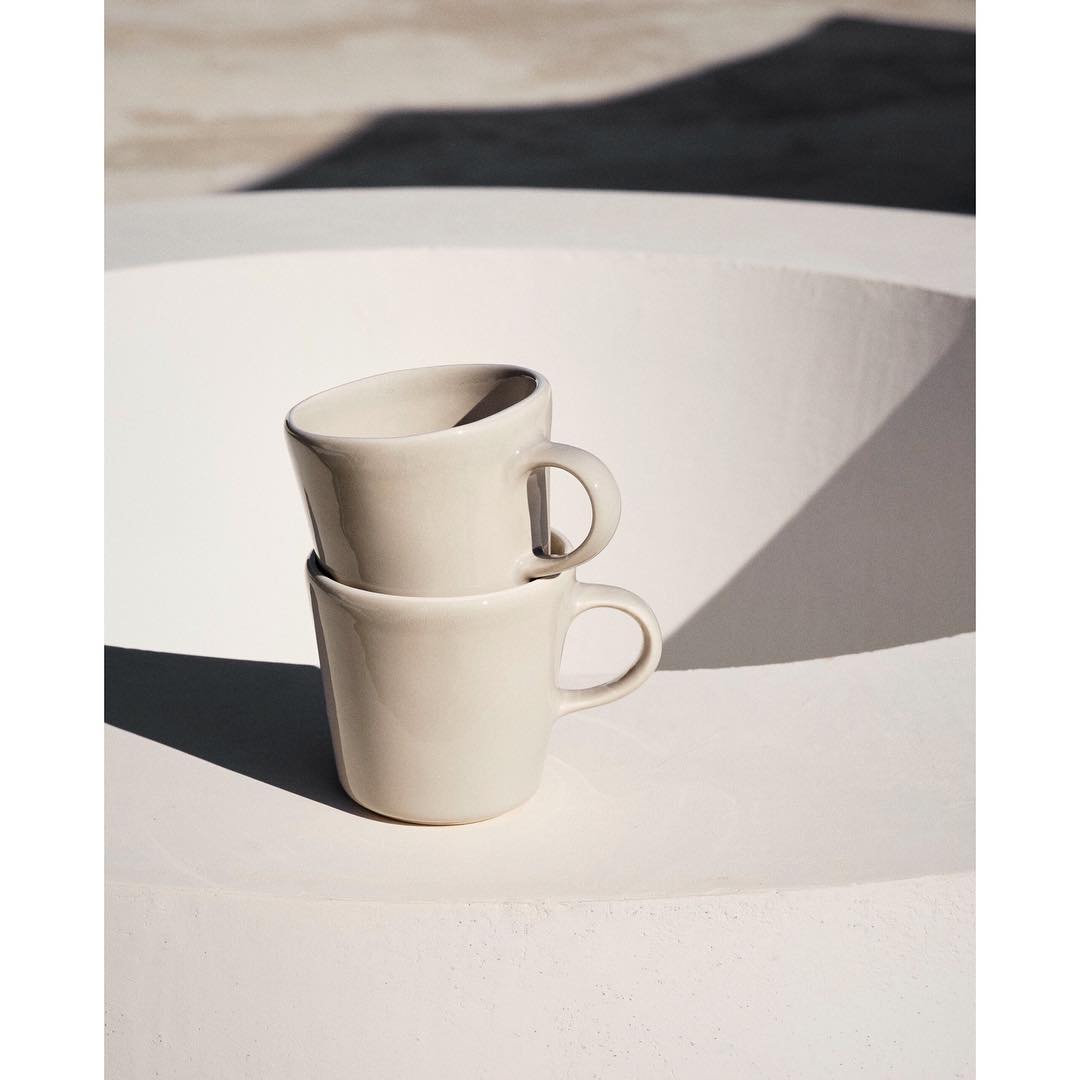 Two ceramic cups stacked on a white surface, bathed in bright sunlight creating stark shadows, with a minimalist and clean aesthetic.