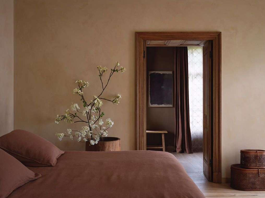 Bedroom interior featuring a bed with brown linens, a flowering branch in a pot, and a doorway leading to another room.